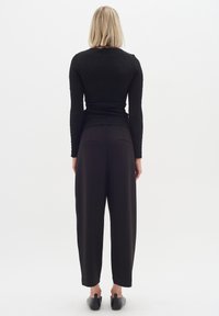 Top lungo a maniche lunghe in tessuto nero abbinato a pantaloni neri a vita alta. L'outfit presenta una vestibilità morbida e dettagli delle cuciture discreti. Scarpe slip-on nere.
