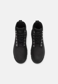 Bottes noires imperméables avec une tige lisse, œillets métalliques, semelle en caoutchouc épaisse et col rembourré. Présente un logo sur la languette.