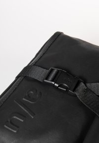 Black fabric bag with a textured surface, featuring a black strap and buckle closure. The brand name is embossed on the material.