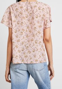 Blusa rosa chiaro con un motivo floreale marrone e blu, maniche corte e dettaglio a chiave sul retro, abbinata a jeans in denim chiaro.