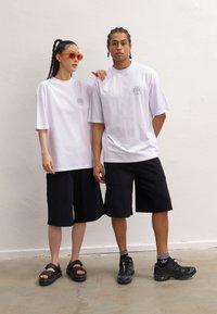 White oversized t-shirts feature a graphic on the chest, paired with black shorts. The female model wears sandals, the male model sports sneakers.