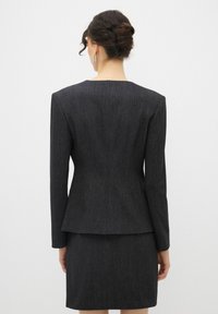 Op maat gemaakte zwarte pak met een aansluitende blazer en kokerrok. De stof heeft een subtiele textuur. Lange mouwen en een ronde halslijn.