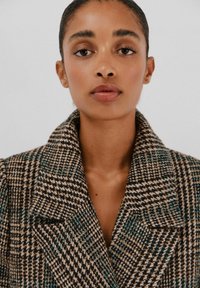 Bruine en zwarte houndstooth jas met teal accenten, grote kraag en gestructureerde pasvorm. De stof heeft een textuur, geweven uitstraling.