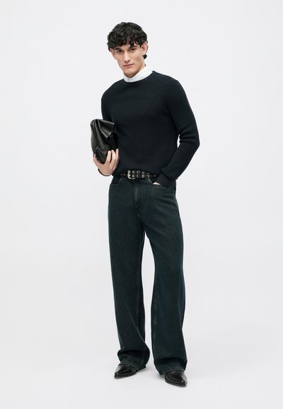 Zwarte trui met textuur, donkerblauwe wijde jeans met hoge taille, zwarte leren clutch en zwarte schoenen met spitse neus.