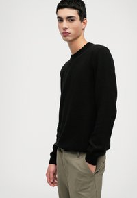 Svart texturerad tröja med rund halsringning och ribbade ärmslut, i kombination med ljusbruna byxor. Modellen står vinklad mot en enfärgad bakgrund.