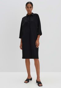 Schwarzes Hemdkleid mit Kragen, Dreiviertelärmeln und gerader Silhouette. Getragen mit schwarzen Slipper-Schuhen. Glatte Stofftextur.
