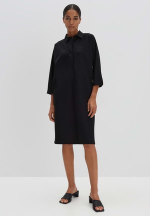 Zwart shirtjurk met een kraag, driekwartmouwen en een rechte snit. Draagt met zwarte instapschoenen. Glad stoftextuur.