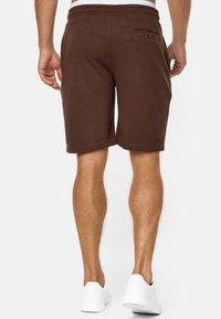 Homme portant un short marron de longueur genou et des baskets blanches, vu de dos contre un fond blanc uni.