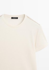 Creme kortærmet t-shirt lavet af blødt stof, med rund halsudskæring og kontrastfarvet brun syning langs skuldrene.