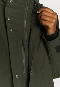 Gros plan d'une main ajustant le col d'une veste vert foncé avec une fermeture éclair, des boutons-pression et un poignet noir visible.