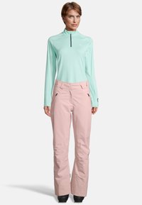 Vrouw in een muntgroene lange mouwen rits-top en lichte roze skibroek met zwarte laarzen, staand tegen een witte achtergrond.