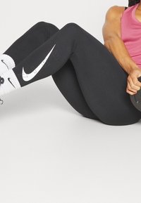 Svarta leggings med en vit Nike-swoosh-logotyp på sidan, kombinerade med vita och svarta mönstrade strumpor. Modellen sitter och håller en hantel.