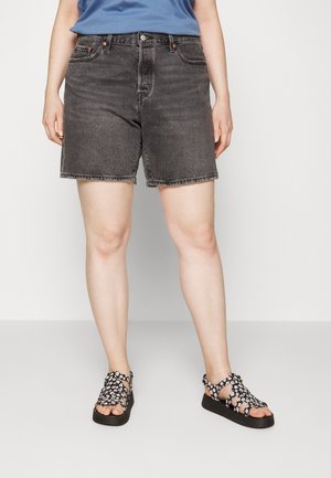 Personne portant un short en jean noir, une chemise bleue et des sandales noires avec des motifs de fleurs blanches, debout devant un fond clair uni.