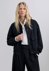 Mujer rubia con pelo ondulado lleva una chaqueta y pantalones de rayas negras sobre una camisa blanca abotonada, posando con una mano en la chaqueta.