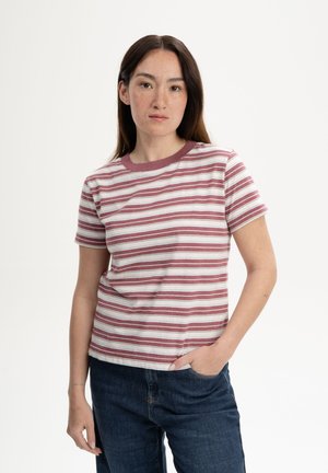 Junge Frau mit langen dunklen Haaren, die ein kurzärmliges, kastanienbraun-weiß gestreiftes Shirt und dunkelblaue Jeans trägt, mit einer Hand in der Tasche.