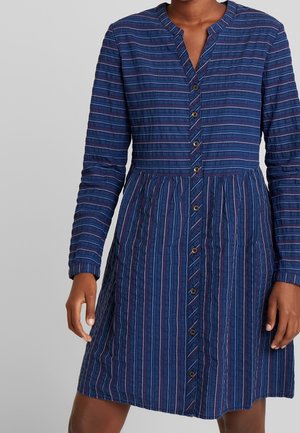 Mujer con un vestido azul de manga larga con patrones de rayas verticales y horizontales y botones en la parte delantera.