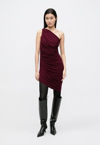 Bordó egyrészes ruha vállnélküli, redőzött textúrával, fekete térdig érő csizmával párosítva. A ruha testhezálló sziluettel és sima anyaggal rendelkezik.