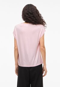 Femme aux cheveux bouclés et foncés, portant un haut à manches courtes rose clair et un pantalon noir ample, debout de dos sur un fond blanc.