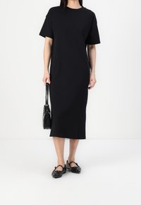 Robe midi noire à manches courtes en coton. Présente une coupe droite, une texture subtile, et est associée à des sabots noirs cloutés et un sac à main.
