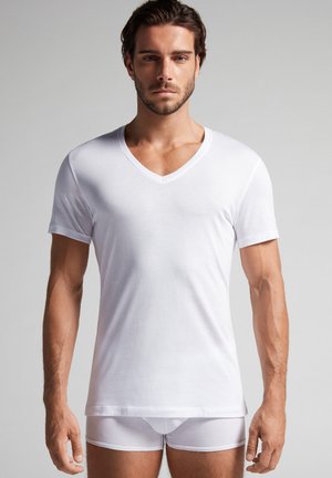 Weißes kurzärmeliges V-Ausschnitt-T-Shirt aus glattem Stoff, kombiniert mit weißer Unterwäsche, getragen von einem Modell vor einem neutralen Hintergrund.