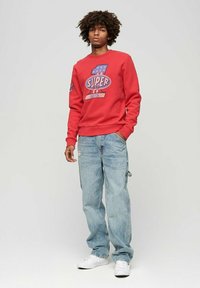 Felpa rossa con stampa grafica, abbinata a jeans baggy azzurri chiari. Il modello indossa sneakers bianche. Design semplice, stile casual.