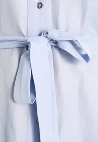 Abito in tessuto azzurro chiaro con cintura in vita, dotato di chiusura a bottoni e una texture liscia. Include un motivo intrecciato.