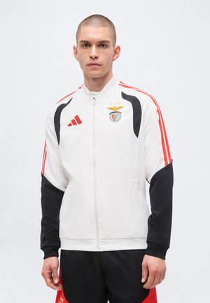 Jeune homme aux cheveux courts portant une veste de sport blanche zippée avec des bandes rouges, des manches noires et un logo du club de football Benfica.