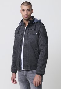 Svart denimjacka med grå huva, dragkedjefickor och knapppuffar. Har ribbade muddar och nederkant, med en strukturerad yta.
