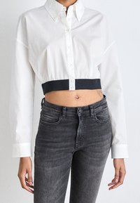 Camicia bianca tagliata con bottoni, con fascia blu navy in vita, maniche lunghe e colletto classico. Abbinata a jeans in denim scuro.