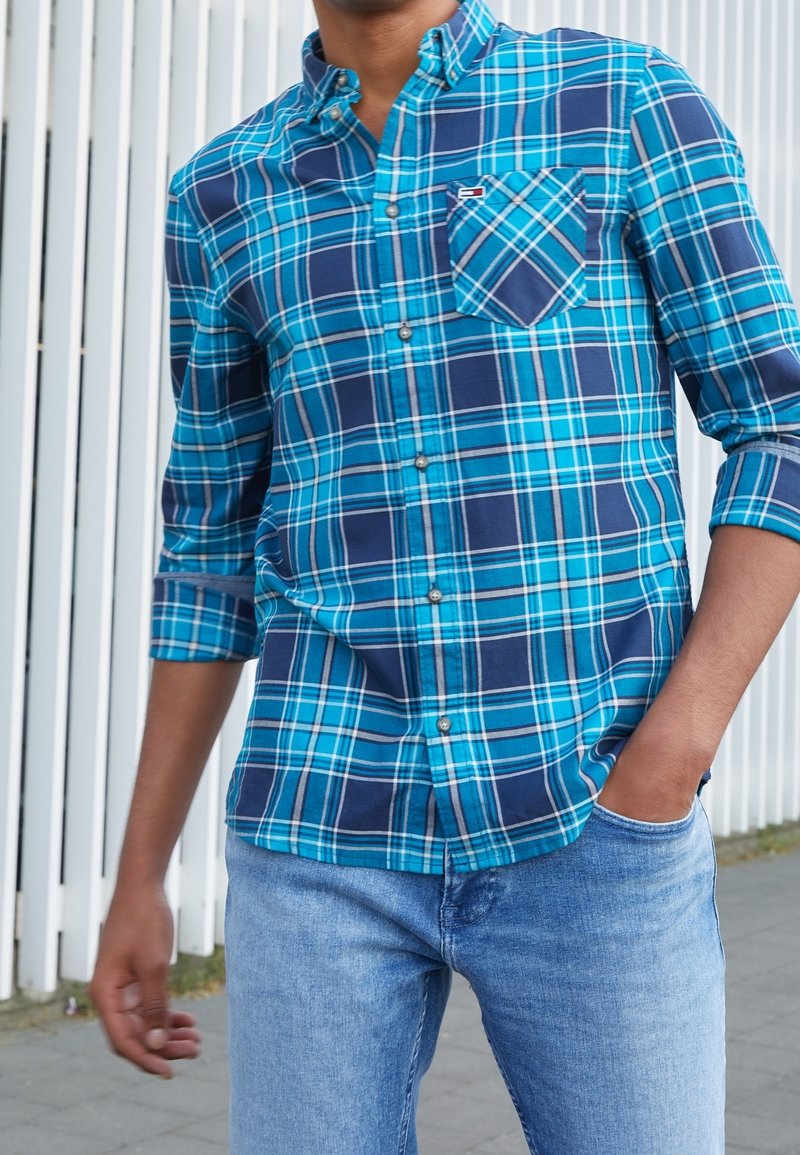 Homme portant une chemise à carreaux bleu et bleu marine à boutons avec les manches retroussées et un jean bleu clair, debout contre une clôture blanche.