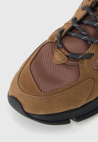 Sapatilhas castanhas com uma mistura de materiais de camurça e malha, com uma superfície texturizada, atacadores escuros e uma sola de borracha preta.