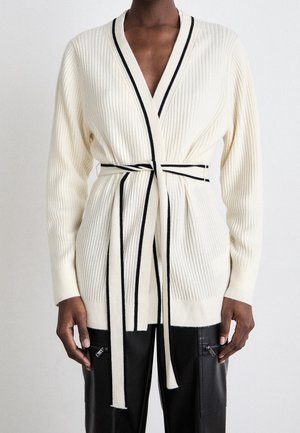 Personne portant un cardigan en tricot côtelé blanc avec des bordures noires et une ceinture assortie, associé à un pantalon en cuir noir avec des poches zippées.