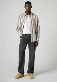 Camicia a righe grigia con bottoni sopra una t-shirt bianca, abbinata a jeans scuri e scarpe slip-on gialle. Il modello è in piedi rivolto verso l'avanti.