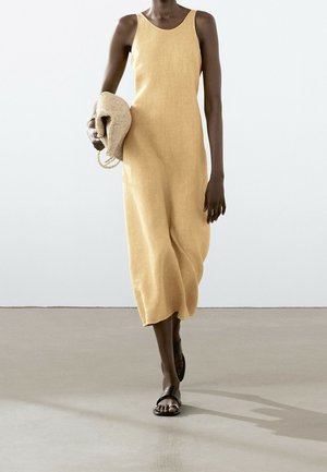 Lniana sukienka bez rękawów w delikatnym żółtym odcieniu, o luźnym kroju i długości do kostek. Modelka trzyma plecioną torbę. Dopełnieniem stylizacji są sandały.