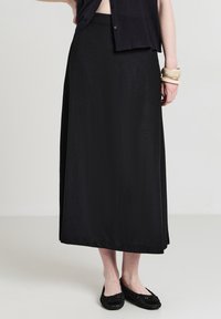 Donna che indossa una gonna midi nera, una maglietta nera con bottoni, delle ballerine intrecciate nere e dei braccialetti beige sul polso destro, in piedi su un pavimento semplice.