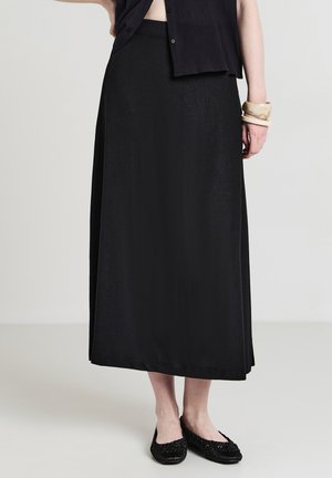 Frau in einem schwarzen Midikleid, schwarzem Knopf-Oberteil, schwarzen gewebten Ballerinas und beigen Armreifen am rechten Handgelenk, die auf einem schlichten Boden steht.