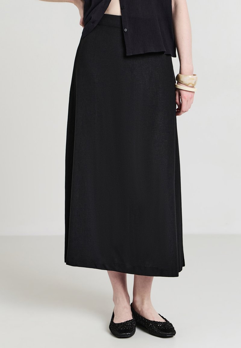 Donna che indossa una gonna midi nera, una maglietta nera con bottoni, delle ballerine intrecciate nere e dei braccialetti beige sul polso destro, in piedi su un pavimento semplice.