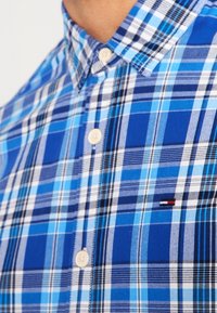 Camicia a quadri blu, bianca e nera con bottoni beige e un piccolo logo ricamato rosso, bianco e blu sul petto.