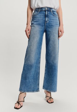 Vrouw in hoge taille, wijduitlopende blauwe denim jeans met een vervaagde wassing en zwarte leren sandalen met hakken tegen een effen achtergrond.