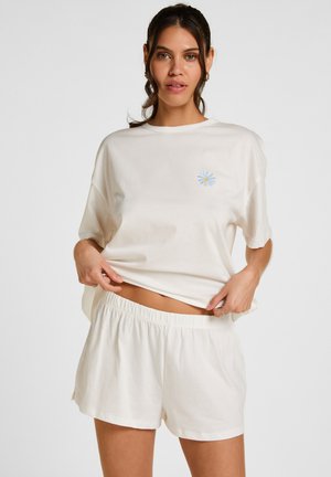 Vrouw draagt loszittend wit T-shirt met klein blauw madeliefjeslogo en bijpassende witte shorts met elastische taille, poseert tegen effen achtergrond.