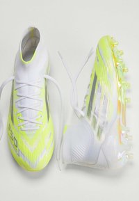 Λευκά και νέον κίτρινα ποδοσφαιρικά παπούτσια με υφή στην επάνω επιφάνεια, λεπτά κορδόνια και διαφανή σόλα με πολύχρωμες τάπες.