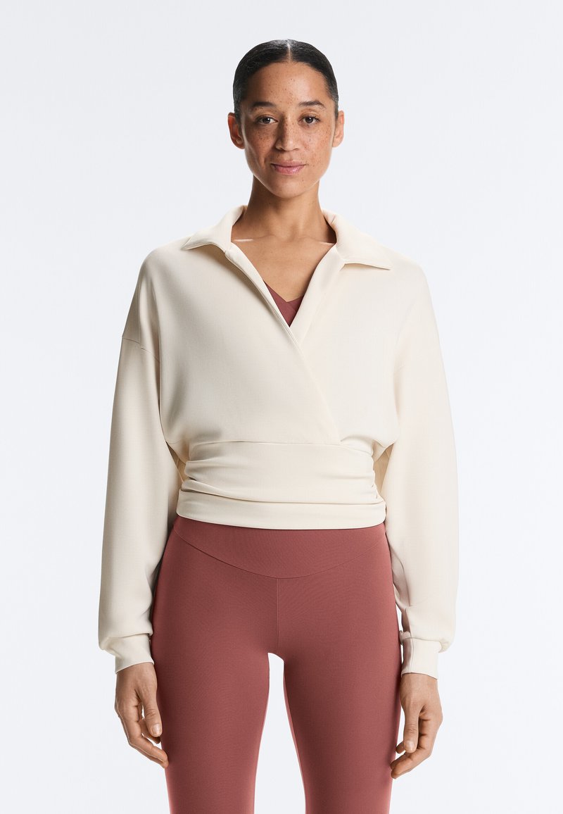 Femme aux cheveux tirés en arrière portant un haut crème à manches longues style cache-cœur et un legging taille haute mauve, debout devant un fond blanc.