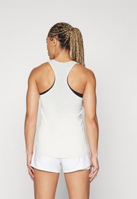 Femme avec des cheveux tressés portant un débardeur blanc et un short de sport blanc, se tenant de dos contre un fond uni.