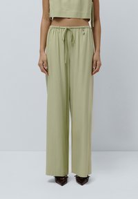 Pantaloni ampi di color verde chiaro, realizzati in un tessuto morbido con cintura elastica e cordoncino, dotati di una piccola accentuazione metallica.