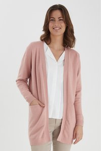 Cardigan rosa chiaro realizzato in materiale morbido, presenta maniche lunghe, un'apertura frontale e due tasche. Indossato sopra una camicia bianca e pantaloni beige.