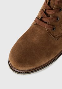 Botte en cuir suédé brun avec un bout arrondi, lacets bruns tressés et coutures visibles sur le bord de la semelle en caoutchouc sur un fond blanc.