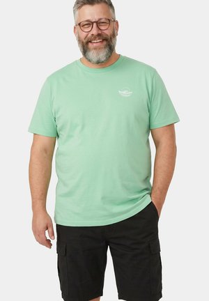 Lächelnder bärtiger Mann mit Brille, einem hellgrünen T-Shirt mit kleinem weißem Logo und schwarzen Cargo-Shorts, steht mit einer Hand in der Tasche.