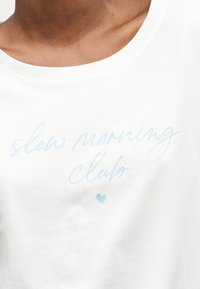 Κοντινό πλάνο ενός λευκού πουκαμίσου με φωτεινά γαλάζια κείμενα με καλλιγραφία «slow morning club» και μια μικρή καρδιά κάτω από το κείμενο.