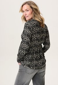 Zwarte top met een wit gespikkeld patroon, lange mouwen en een geplooide taille; gecombineerd met grijze jeans. Zachte stoftextuur.