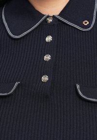 Marineblå ribstrikket polo med krave, sølvfarvede knaplukninger og kontrasterende hvid piping på kraven og lommekanterne.